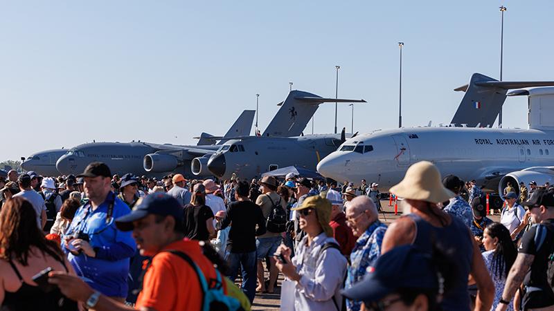 Crowd of visitors walking along an airfield apron with several Royal Australian Air Force transport aircraft parked in the background during a public open day, viewed under clear daytime skies.