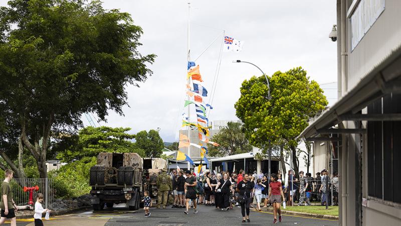 Crowds of visitors gather along a roadway at a military open day or public event. A tall mast in the centre of the scene is decorated with colourful maritime signal flags and a white ensign-style flag flying at the top.