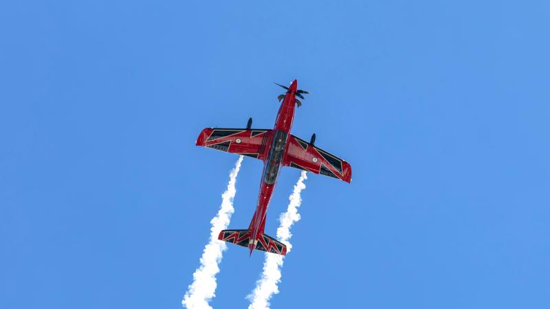 Underside view of a red aerobatic aircraft flying overhead against a clear blue sky. White smoke trails stream from both wings, creating two vertical plumes behind the aircraft. The plane features a bold red, black, and white livery, and is centered in the frame with extensive blue sky surrounding it.